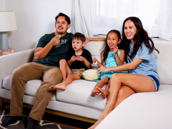 Father, son, daughter and mother watching tv while sitting on the couch and eating popcorn.